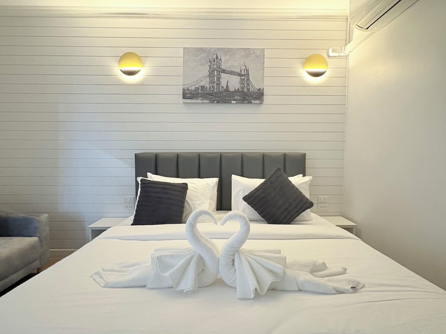 Ett hotellrum med en säng, två vägglampor, en inramad bild av en bro och en konstnärligt vikt handduk som pryder sängen.