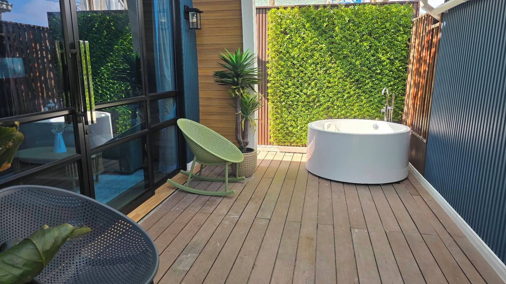 Una terraza de madera con un jacuzzi, una mecedora verde y un ventanal que da a una habitación.