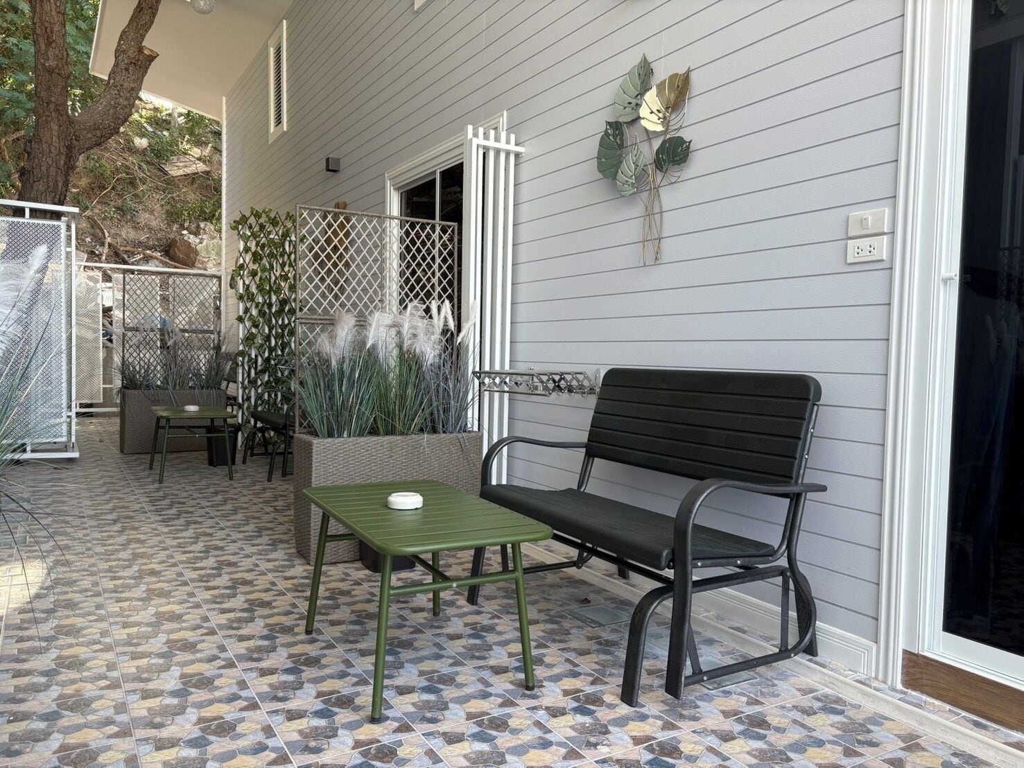 Eine Terrasse mit einem grünen Tisch, einer schwarzen Bank, einem weißen Zaun und einem Pflanzgefäß mit hohen Pflanzen.