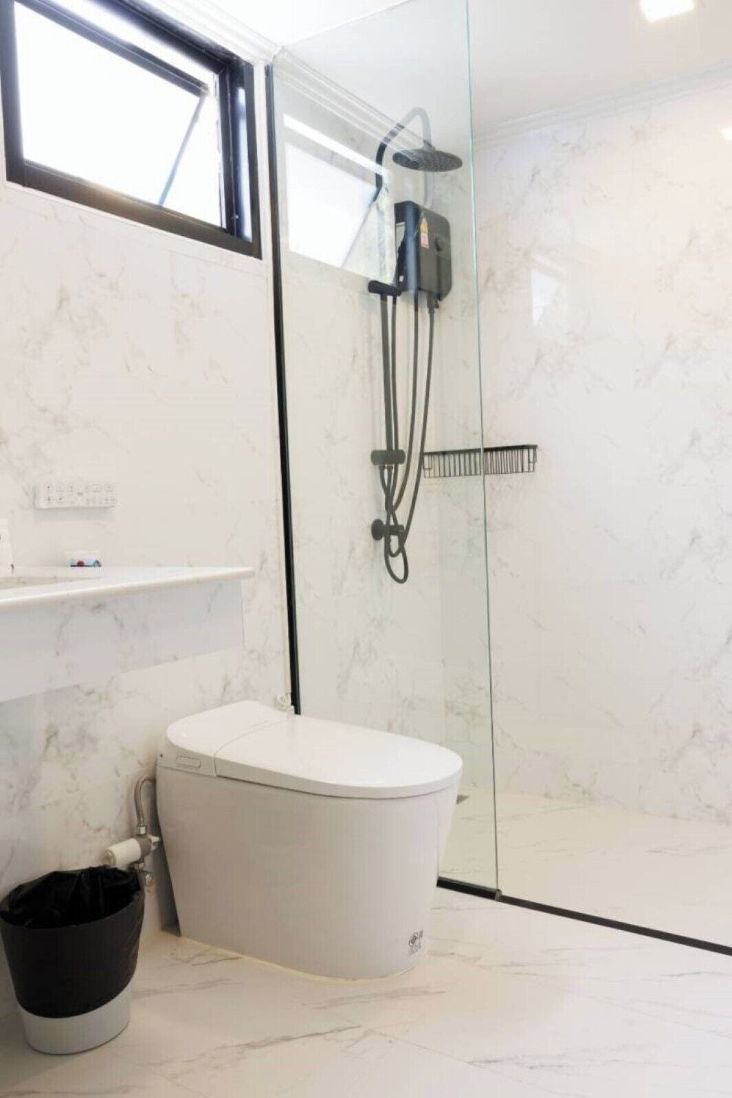 A bathroom with a white toilet, a black trash bin, a glass-enclosed shower area, and a marble-tiled wall.