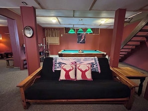 Ein Zimmer mit Billardtisch, einer Couch mit Dekokissen, einer Uhr und einer Treppe.
