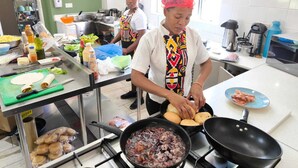 Une cuisine dotée de plans de travail en acier inoxydable, d’une cuisinière et de divers ustensiles de cuisine. On y voit deux chefs portant des tabliers colorés et des couvre-chefs.