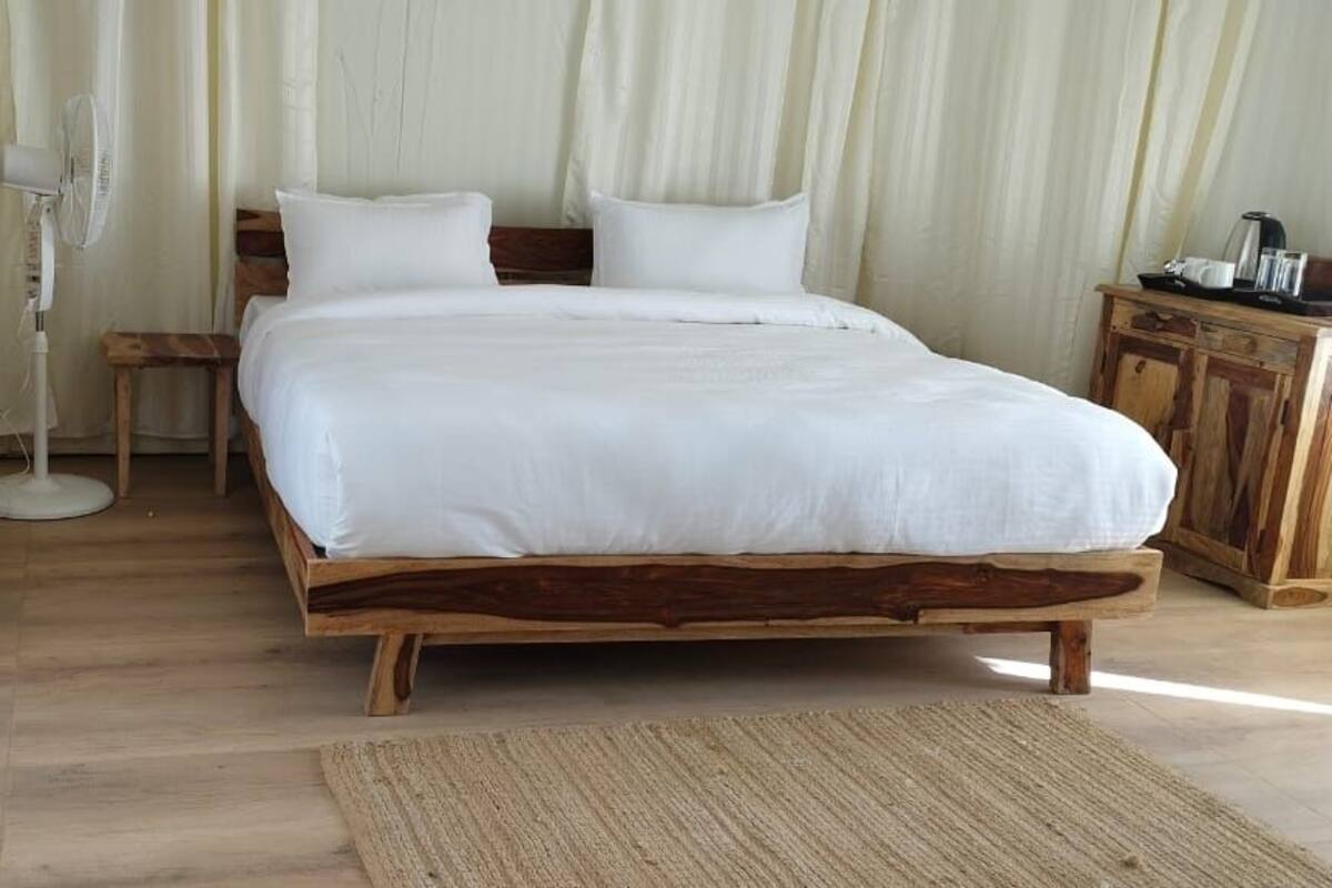 Un lit en bois avec une literie blanche, dans une pièce au plancher de bois et dotée d’une grande fenêtre avec des rideaux.