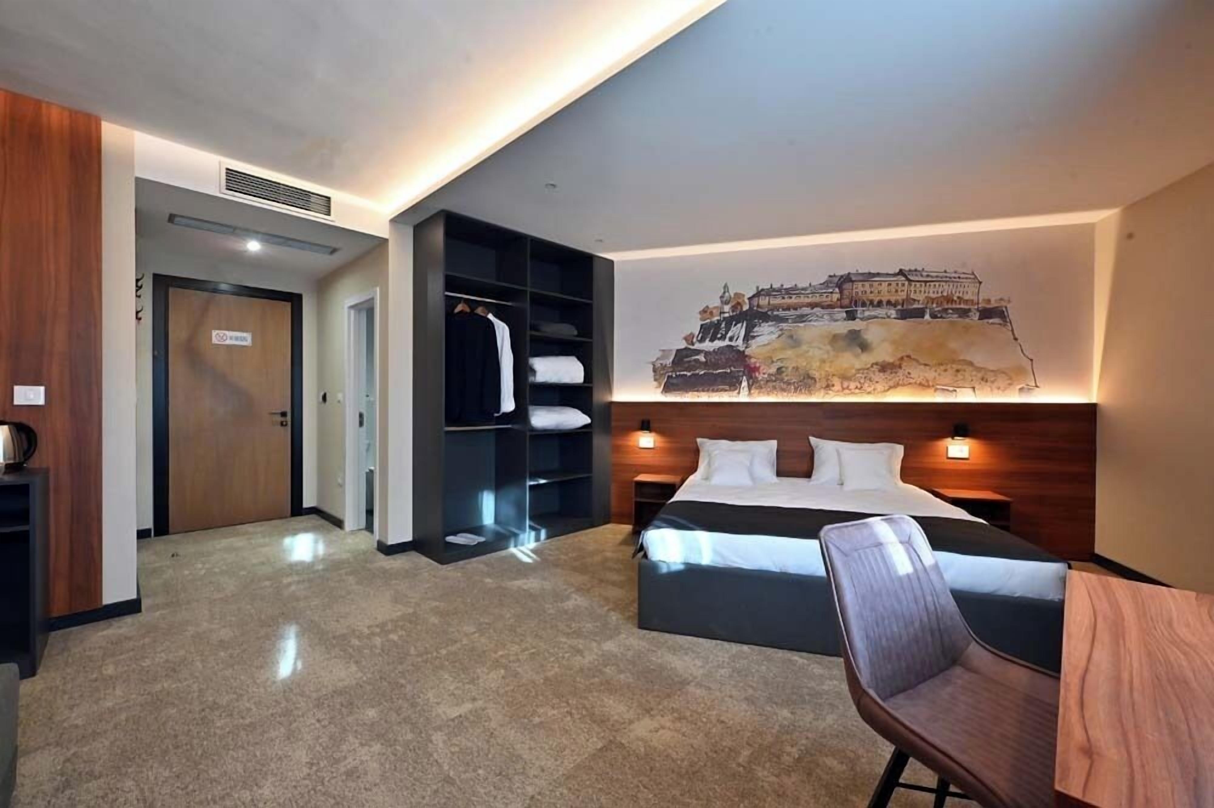 A hotel room with a bed, a chair, a desk, a wardrobe, and a painting on the wall.