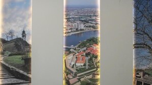 Une pièce avec deux fauteuils verts et une petite table en bois, un grand tableau vertical représentant un paysage urbain, et une vue sur un pont.
