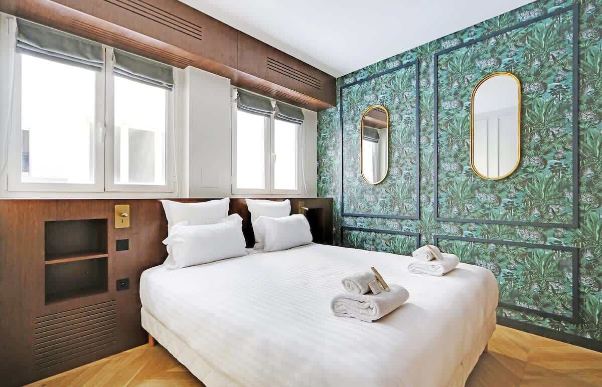 Hotelkamer met een groot bed, twee ramen, een houten hoofdeinde en een groene, patroonbehangen muur met twee spiegels.