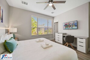 Una habitación de hotel moderna con una cama grande, un escritorio, una silla, un ventilador de techo, un televisor y una ventana con persianas.