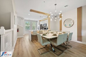 Un comedor moderno con una mesa grande dispuesta para seis personas, sillas azul claro, una araña y una cocina al fondo.