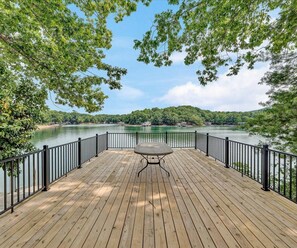 Una terraza de madera con una mesa y barandilla, con vista a un lago, árboles y edificios al fondo.