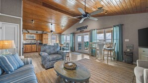 A spacious living area with a sofa, coffee table, and a dining table with chairs. A kitchen is visible in the background, and a large window offers a view of the beach.