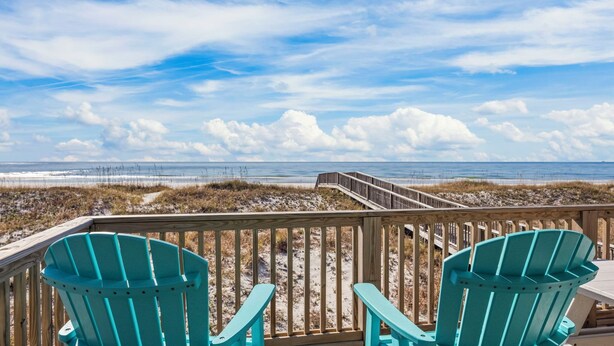 A wooden deck with turquoise Adirondack chairs overlooking a beach with a wooden boardwalk and a view of the ocean.