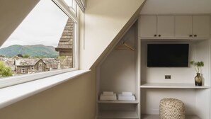 Una habitación con techo inclinado, un televisor montado en la pared, un perchero colgante, un estante pequeño con toallas y vistas a edificios y montañas a través de una ventana.