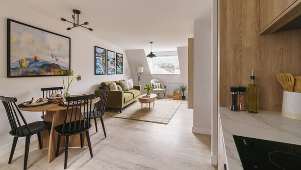 Una sala de estar moderna con comedor, que incluye una mesa de comedor de madera, sillas negras, un sofá y una mesa de centro. Hay obras de arte enmarcadas en las paredes y un ventanal que proporciona luz natural.