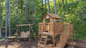 Una casita de juegos de madera con un columpio y un banco en un bosque.