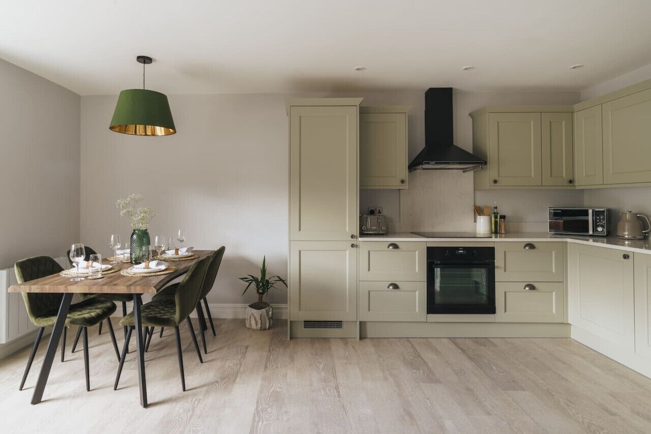 A modern kitchen with light green cabinets, a black range hood, and a dining area with a wooden table and chairs.