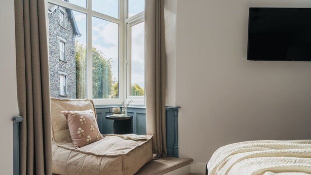 A bedroom with a window seat, a bed, a small table, and a TV mounted on the wall.