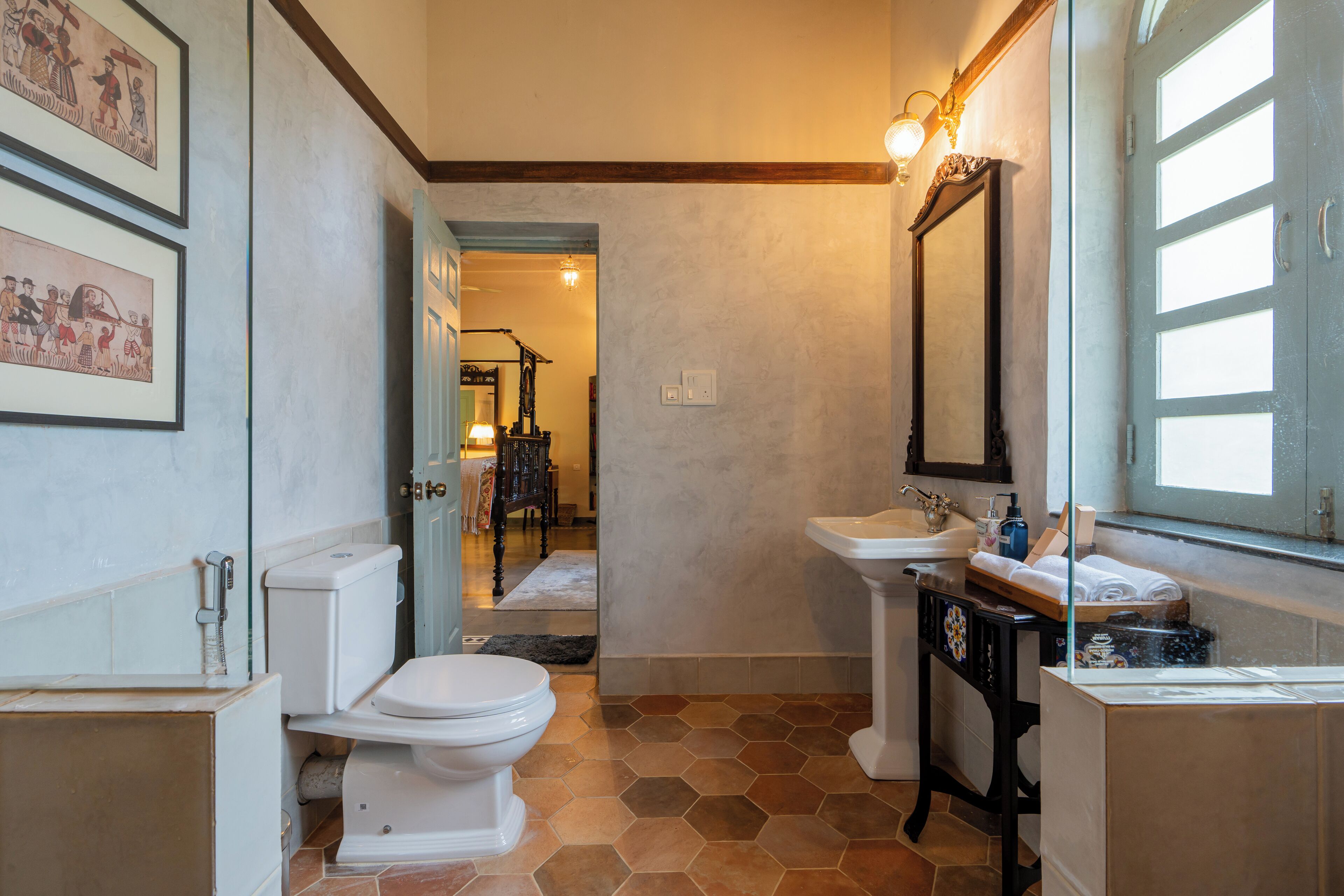 A bathroom with a toilet, sink, and mirror. There are framed pictures on the wall and a window with blinds.