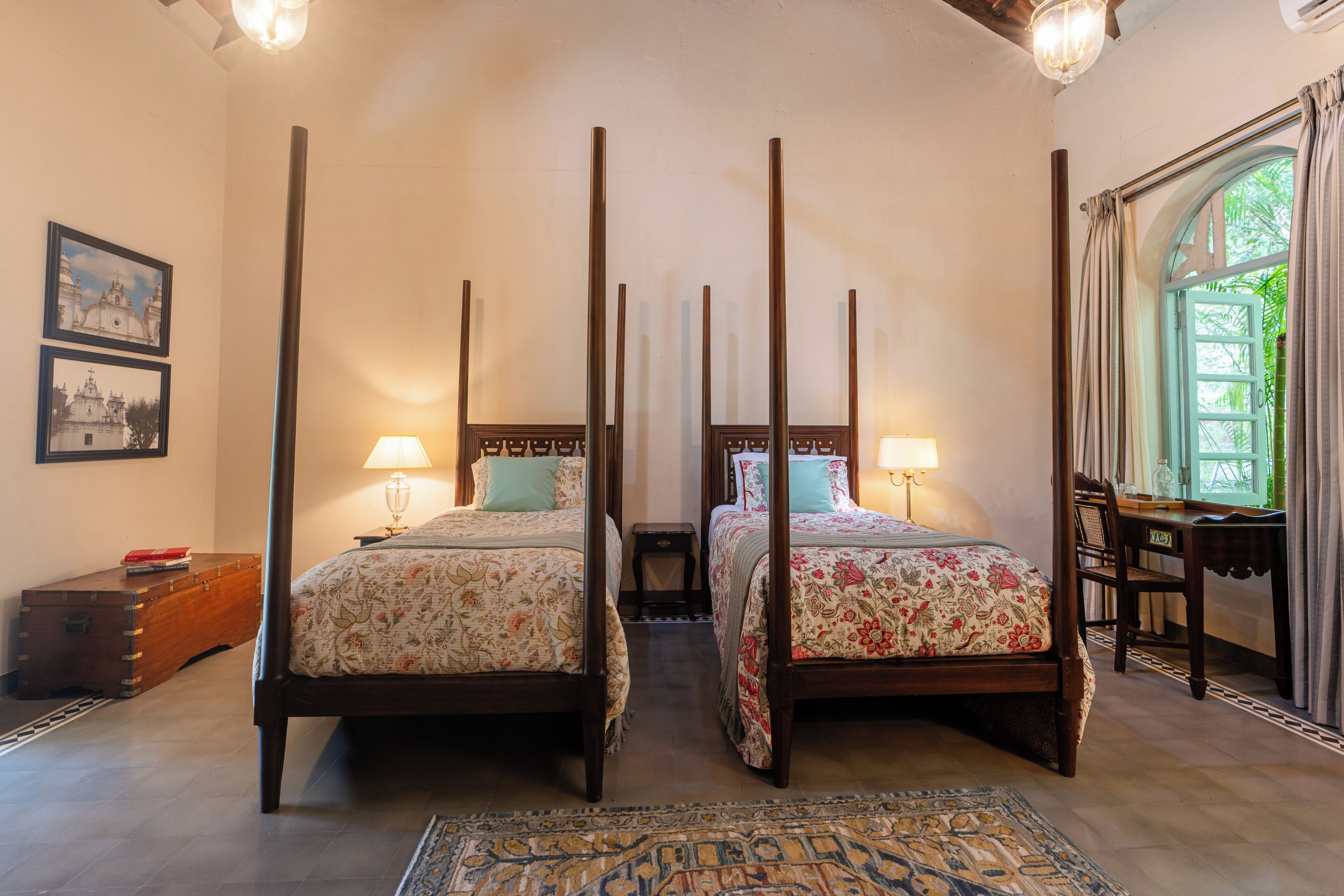 A four-poster bed with floral bedding, a wooden nightstand, a desk with a chair, and a large window with curtains.