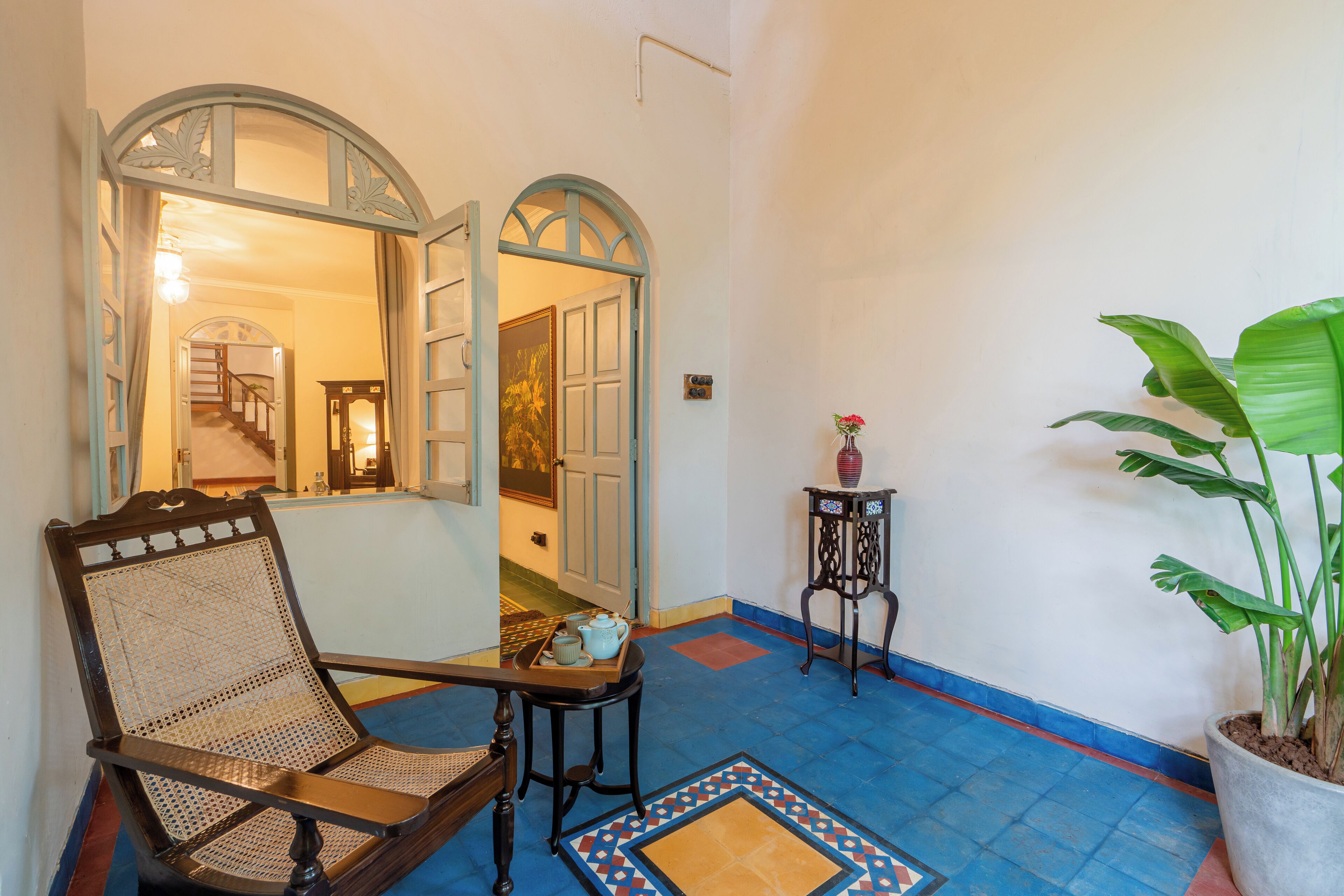 A room with a wooden chair, a small table with a tray, a potted plant, and a tiled floor.
