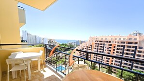 Un balcon avec une table en bois et des chaises blanches, donnant sur un quartier résidentiel et un plan d’eau.