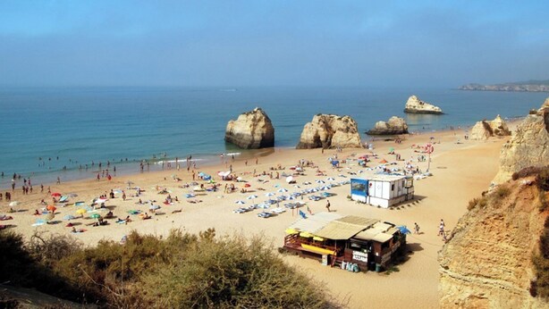 Une plage avec de grandes formations rocheuses, une étendue de sable et plusieurs parasols de plage.