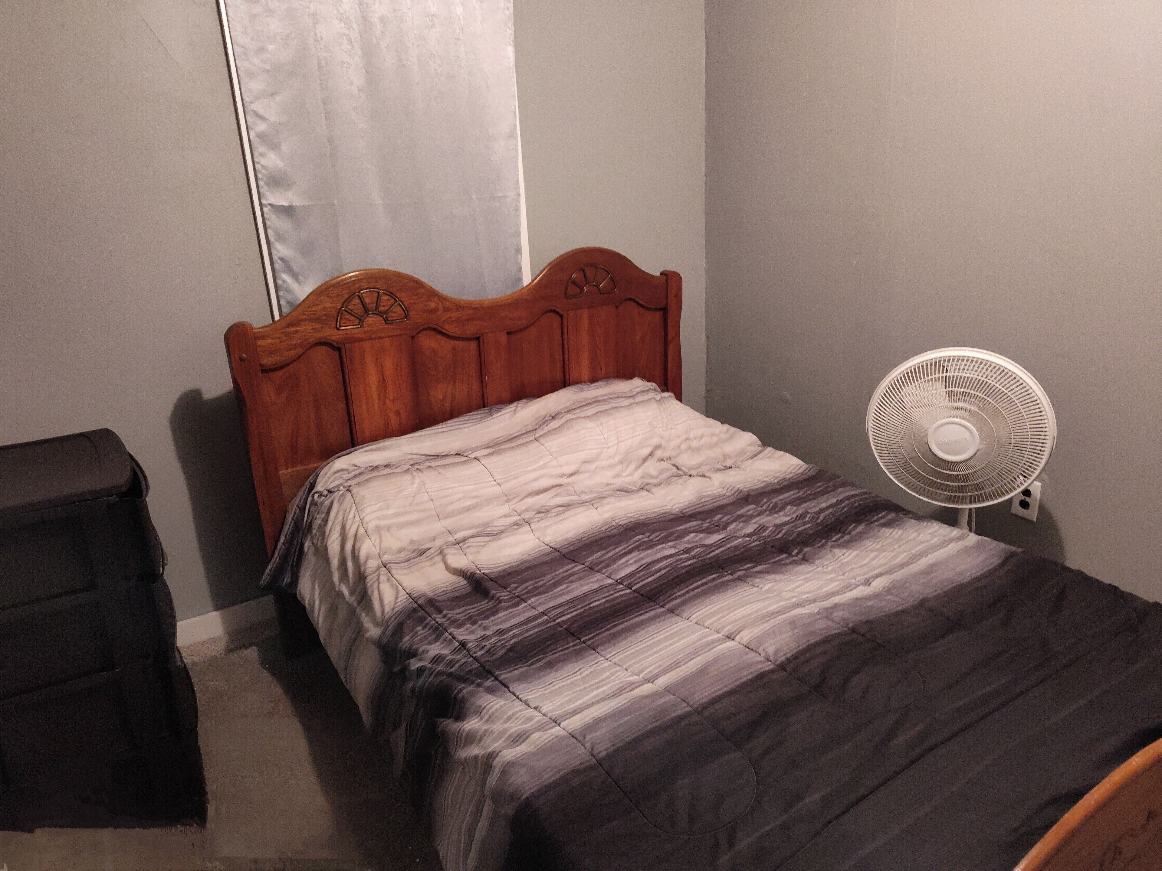 A bedroom with a wooden bed frame, a white wall, and a standing fan.