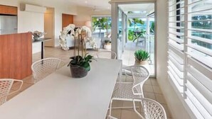 Uma área de jantar moderna com uma mesa e cadeiras brancas, uma planta em vaso e vista para o exterior através de grandes janelas.