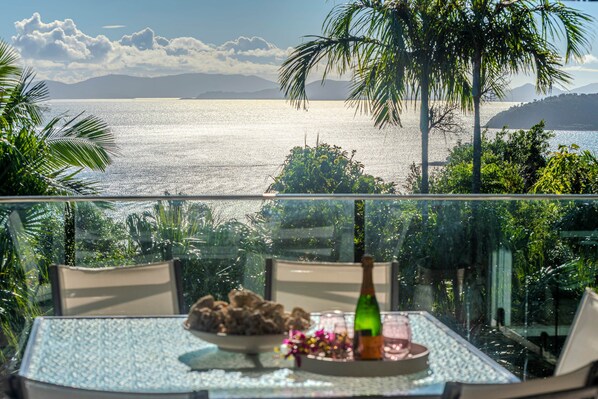 Ein Tisch mit einer Flasche, einer Schüssel und einem Glas auf einem Balkon mit Blick auf ein Gewässer, Palmen und Berge in der Ferne.