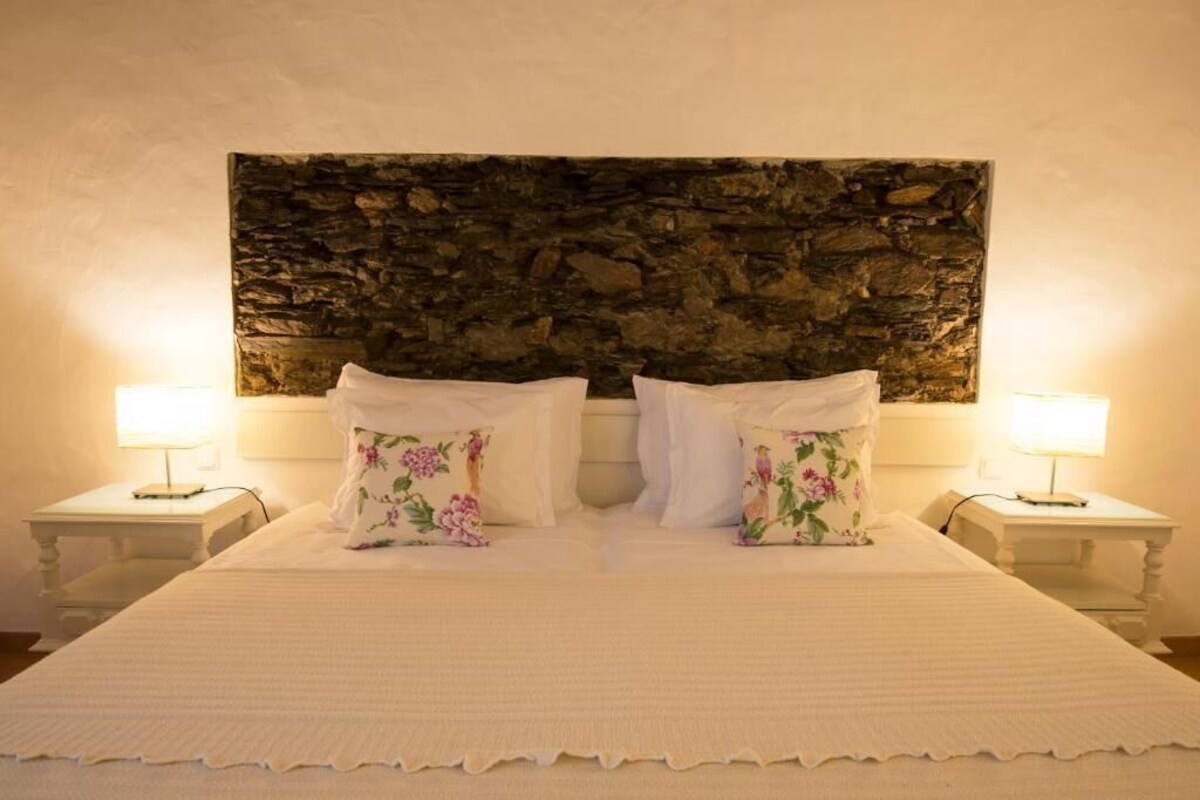 Una cama con cabecera de piedra, dos mesitas de noche con lámparas y almohadas estampadas.