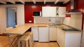 Een kleine keuken met een magnetron, fornuis, spoelbak en een compacte koelkast. Er is een houten tafel met stoelen en een deur naar een andere kamer.
