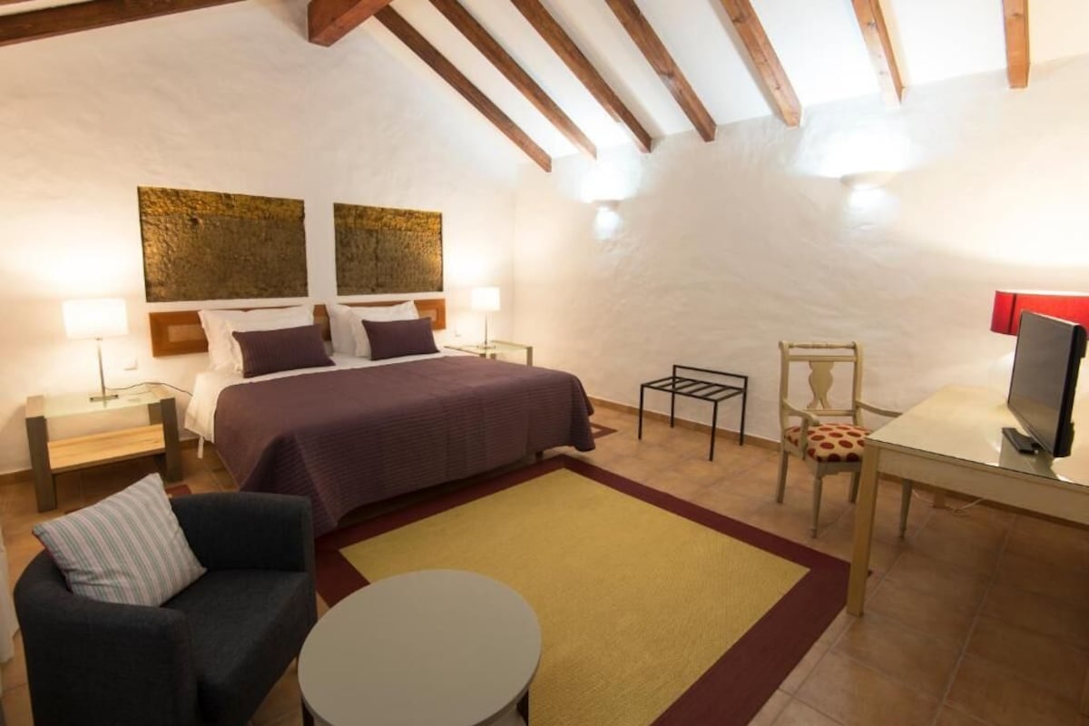 Un dormitorio con una cama, dos mesitas de noche, una silla, una mesita pequeña y un televisor.