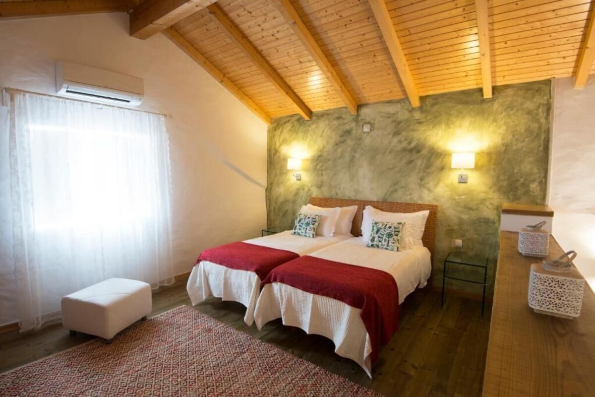 Un dormitorio con dos camas, techo de madera, pared verde con textura y una pequeña mesita de noche con lámpara.