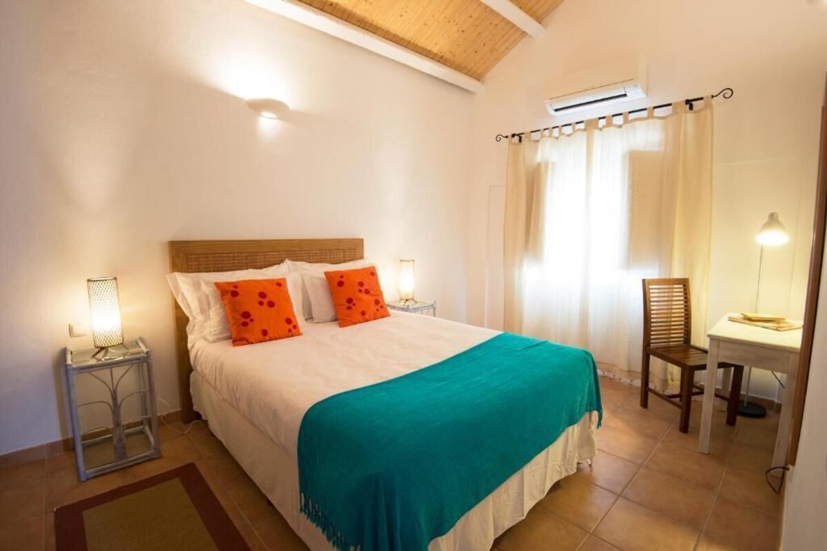 Un dormitorio con cama, mesita de noche, silla, escritorio y ventana con cortinas.