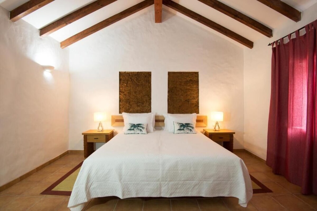 Un dormitorio con una cama grande, vigas de madera en el techo, dos mesitas de noche con lámparas y una cortina roja.
