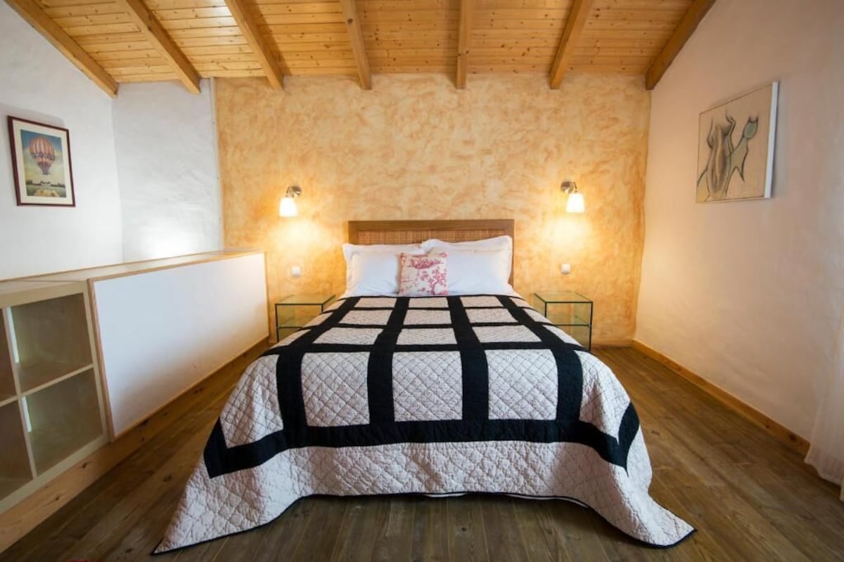Un dormitorio con techo de madera, colcha acolchada, dos lámparas de noche y un cuadro colgado en la pared.