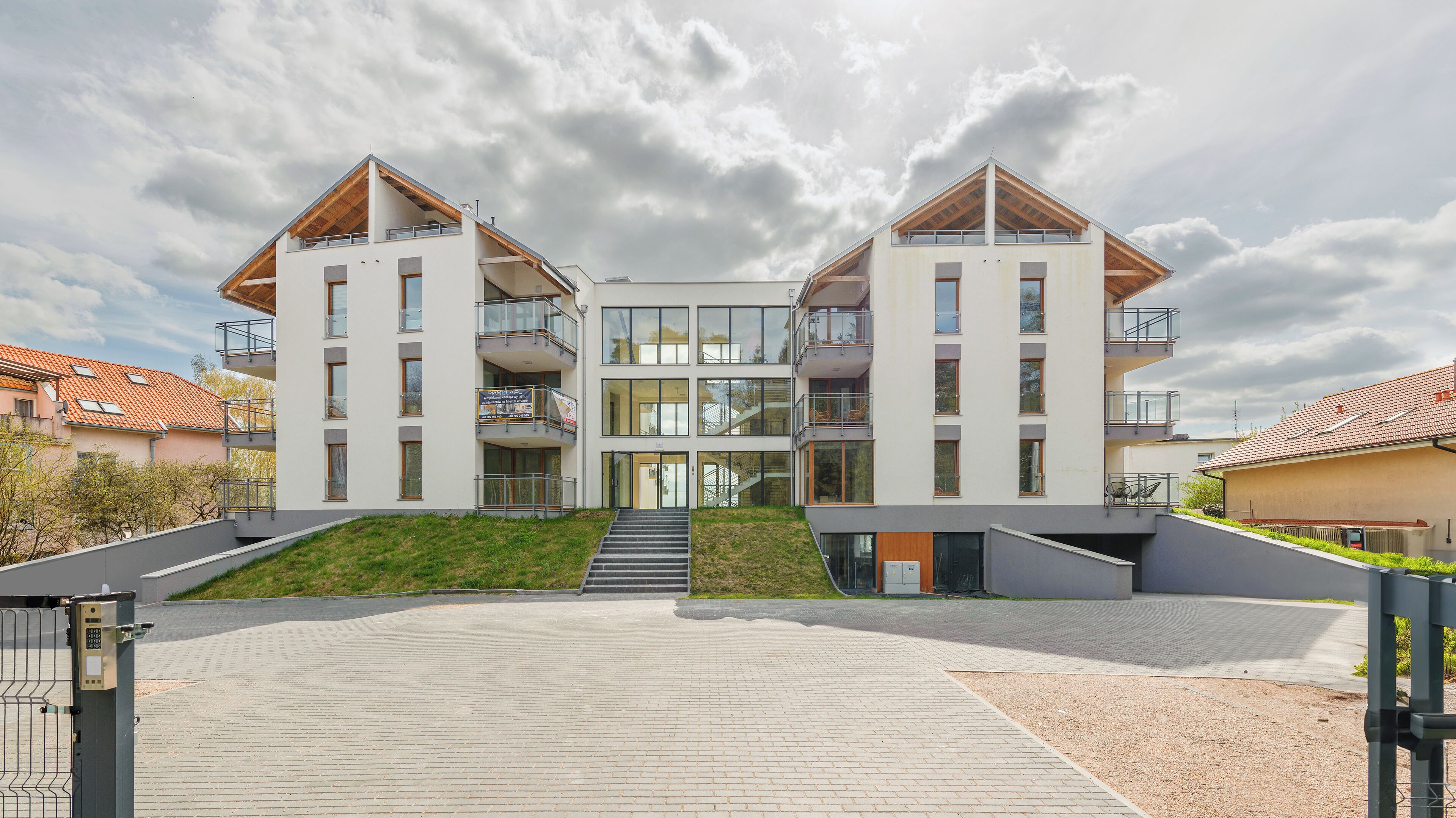 A modern multi-story residential building with balconies, a central staircase, and a paved entrance area.