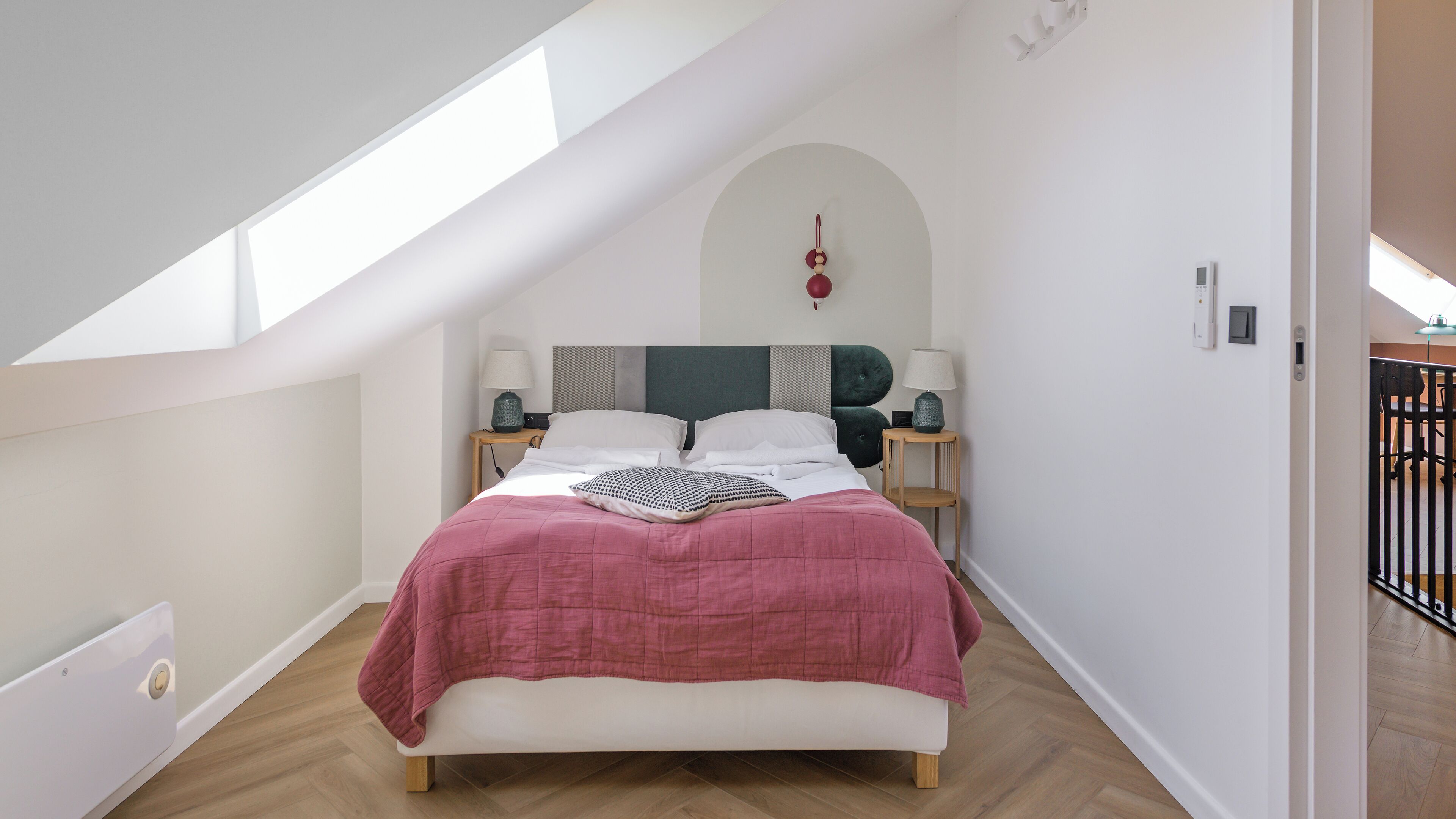 A bedroom with a sloped ceiling, a bed with a pink bedspread, two bedside lamps, and a wall-mounted light fixture.