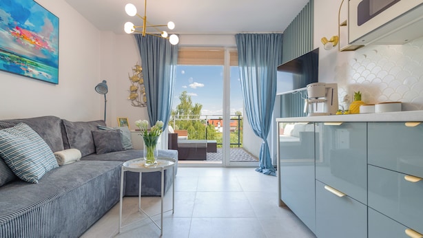 En modern lägenhet med en grå soffa, ett litet sidobord med en vas med blommor, en balkong med utsikt över staden och ett kök med en kaffemaskin.