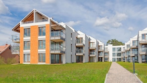 A modern multi-story residential building with balconies, large windows, and a paved pathway leading to the entrance.
