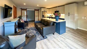 A modern living room with a sofa, a coffee table, and a TV. There is a kitchen area with cabinets and a sink.