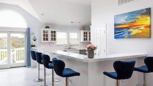 A modern kitchen with white cabinets, a central island with bar stools, and a large window overlooking a balcony. - Whalehead Adventure With Ocean Views, Pool, Spa (Corolla)