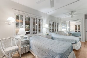 Ein Schlafzimmer mit zwei Betten, einem Korbstuhl, einem Nachttisch mit Lampe und einem großen Fenster mit Rollläden.