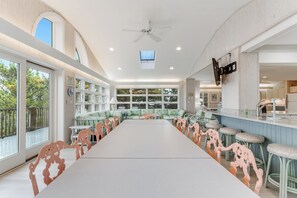Ein geräumiges Zimmer mit einem langen Tisch, grünen Polsterstühlen, einem Deckenventilator und großen Fenstern mit Blick auf Bäume.