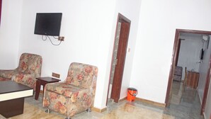 A living room with two floral armchairs, a coffee table, a flat-screen TV mounted on the wall, and a wooden door leading to another room.