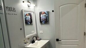 Un baño con inodoro, lavamanos y espejo. Hay luces empotradas en la pared y una puerta.