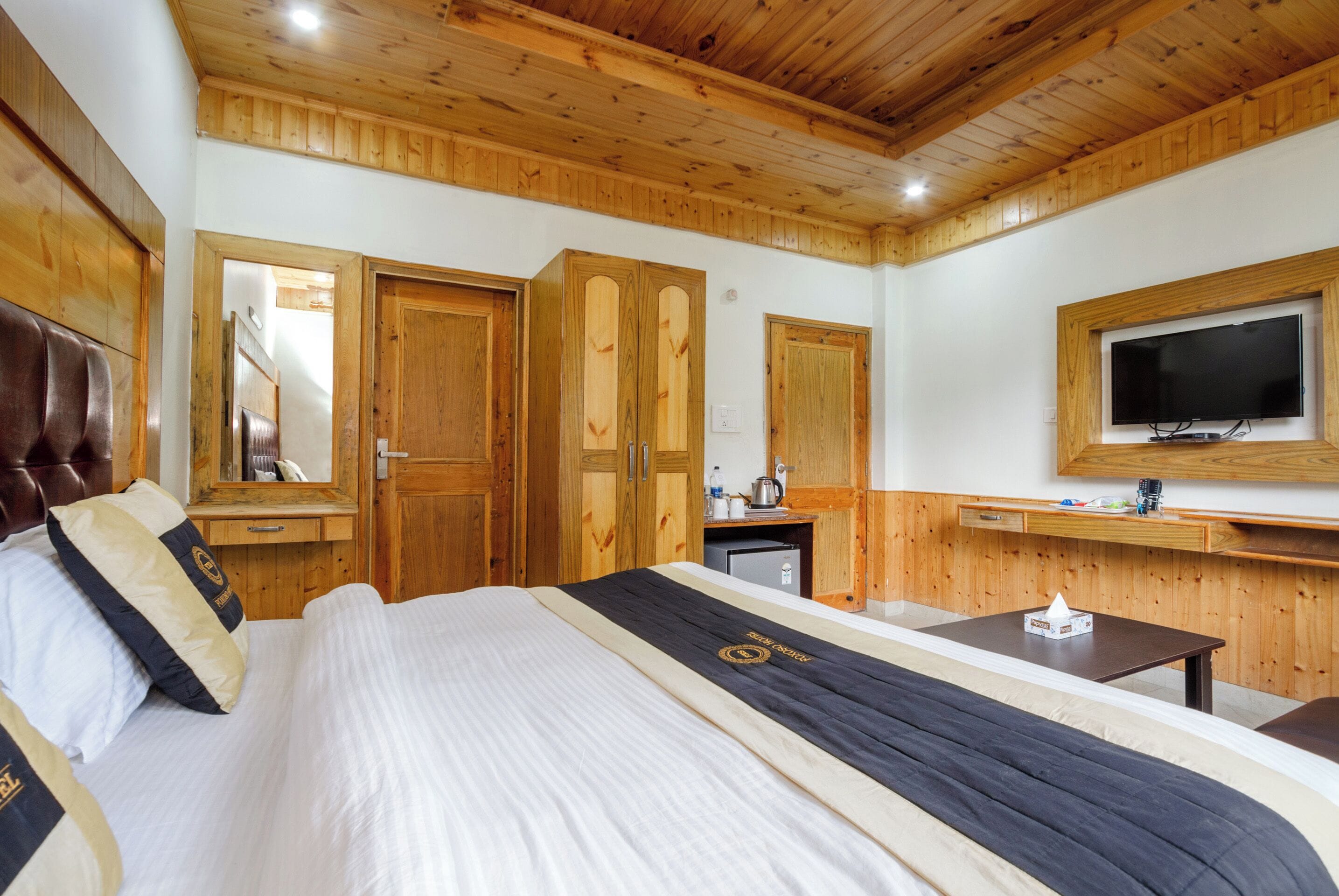 A hotel room with a wooden ceiling, a bed with a black and white bedspread, a wooden wardrobe, a small desk with a television above it, and a mirror on the wall.