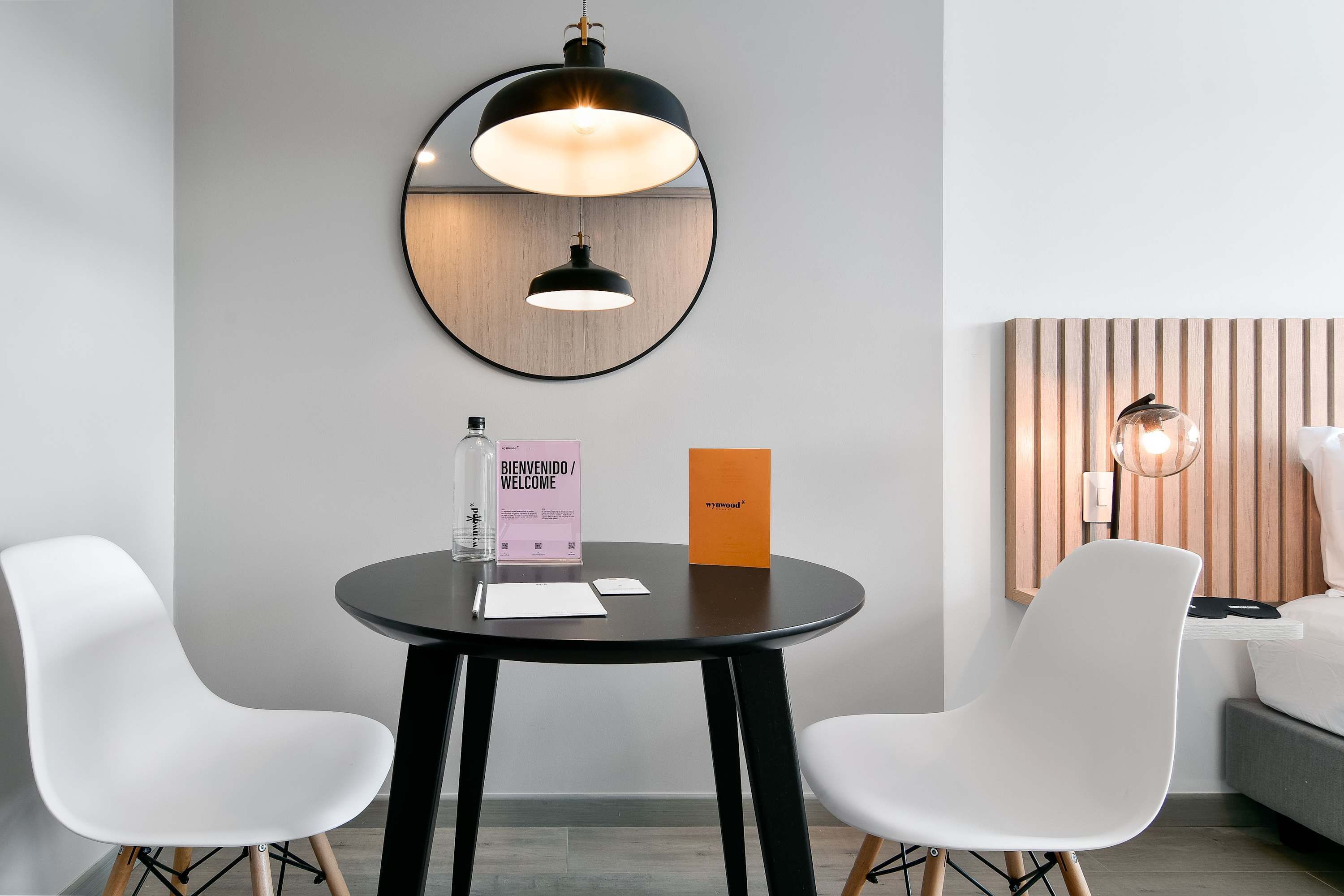 Una habitación de hotel moderna con una mesa redonda, dos sillas blancas, un espejo redondo, un cabecero de madera con listones y una lámpara de noche.