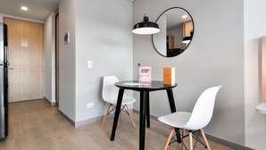 Une chambre d’hôtel moderne avec une table ronde, deux chaises blanches, un luminaire suspendu noir, un miroir rond et une porte en bois.
