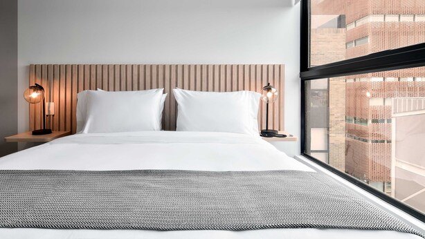 Une chambre d’hôtel moderne avec un grand lit, deux lampes de chevet, une vue sur un immeuble en briques par la fenêtre et un tapis à motifs sur le sol.