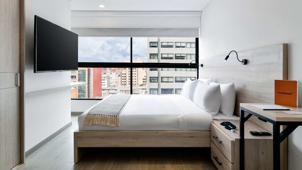 A modern hotel room with a large bed, a desk, a flat-screen TV, and a view of the cityscape.
